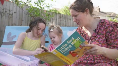 Three people sit together in what looks like a garden or backyard space. An adult is holding a “Little Red Riding Hood – Phonics Story Book”, reading it aloud. Two children sit close by: one is watching the book with interest, while the other is focused on a small container filled with craft or learning materials. They’re gathered around a low wooden table, and behind them is a painted bench decorated with playful animal illustrations — squirrels, trees, and bright colours. A wooden fence and leafy trees frame the scene