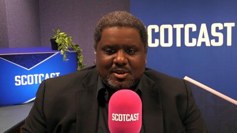 A man with short afro hair and a goatee wearing a black suit jacket and black shirt talking into a Scotcast-branded mic