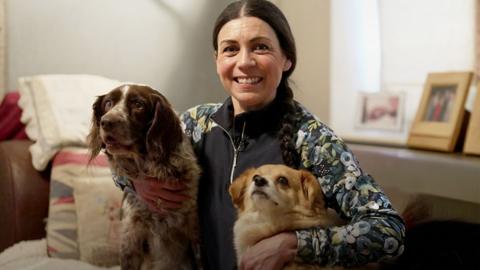 Helene Svinos sits on a sofa in between two dogs, with her arms around them. She is smiling and wearing a dark gilet over a patterned top, and leggings, with her dark hair tied back. There are cushions on the sofa and photo frames in the background.