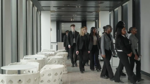 Students walk into the office which is sleek and corporate. They are dressed smartly. White tables and chairs can be seen to the left.