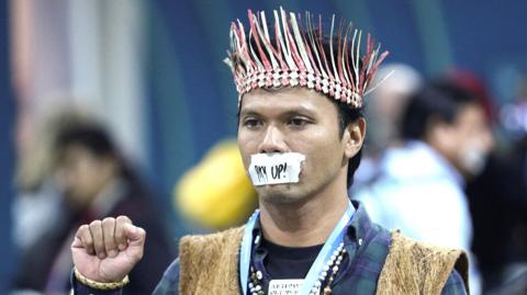 An environmental activist wearing a woven headdress holds his fist up with a piece of tape over his mouth with "pay up!" written on it