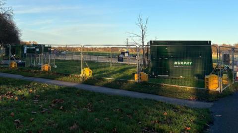 A large green tank stands at corner of green field surrounded by metal fences in a park.