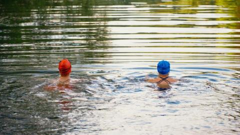 Two female open water wild swimmers in a lake are wearing orange and blue swimming caps. They are facing away from the camera and appear to be swimming breaststroke side by side with their heads out of the water. The water looks still and you can see the reflection of greenery in the distance.