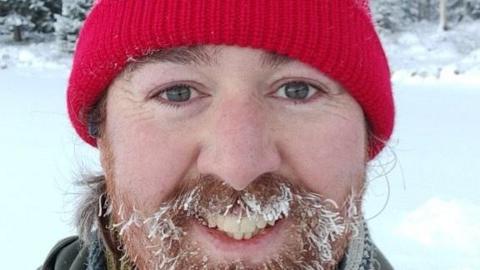 Oliver Eagle-Wilsher is smiling at the camera. He has a red beanie hat on. He is stood outside in the snow and has snowflakes on his ginger beard and hair. 