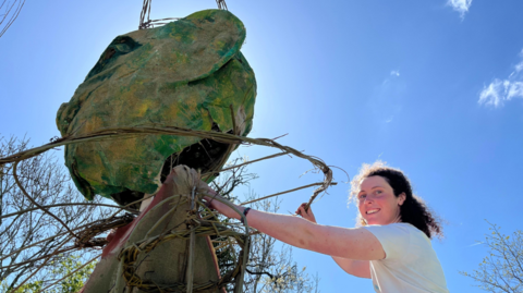 A woman stands outside next to a large puppet with a green head. It has rope around it.