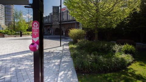 A small circular bin attached to a black lamppost. There is a sign above it saying "gum here" with an arrow pointing down to the bin. 