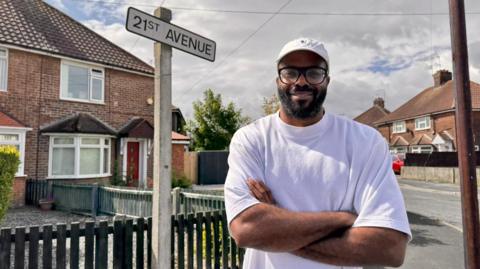 A man wearing a white cap, black rimmed glasses and a white t-shirt stands with his arms folded on a street corner. He is standing in front of a road sign which reads "21st Avenue" and a number of red brick houses are visible in the background.