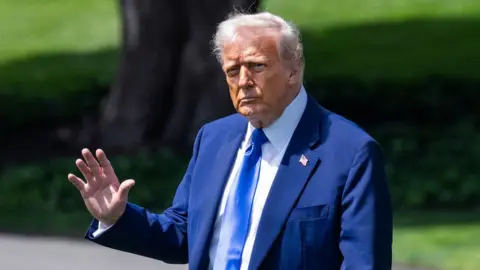 Donald Trump wearing a navy suit, blue tie and white shirt, holding his hand up in a wave