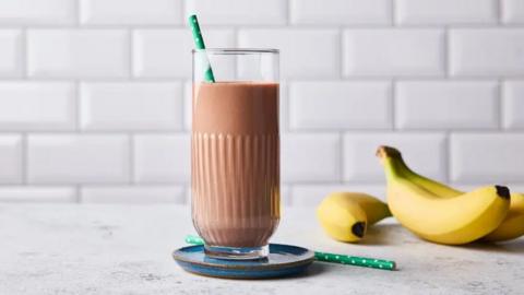 A glass full of chocolate milk with a straw in it. to the right in the background are three bananas.