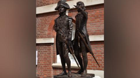 A statue features two figures both in uniform. The statue stands in front of a red brick building with an arched window