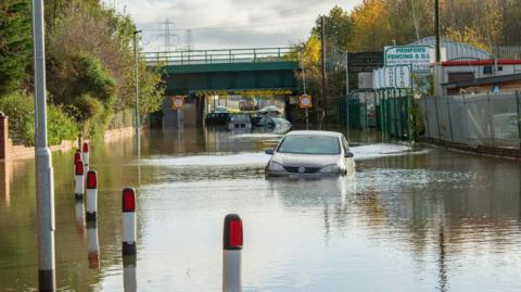 Cars, covered by water up to their lights, sit in flood waters on a road also obscured by flooding.