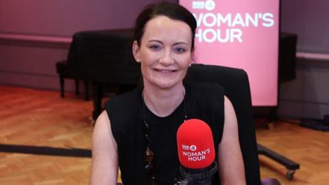 Head and shoulders of Dr Helen Wall sitting behind a red microphone in the Women’s Hour studio. She is wearing a black sleeveless top and she is smiling. 