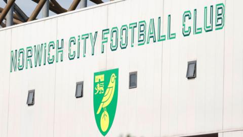 Green writing saying "Norwich City Football Club" on a white external wall of the Carrow Road stadium above the club's logo of yellow canary on a green shield.
