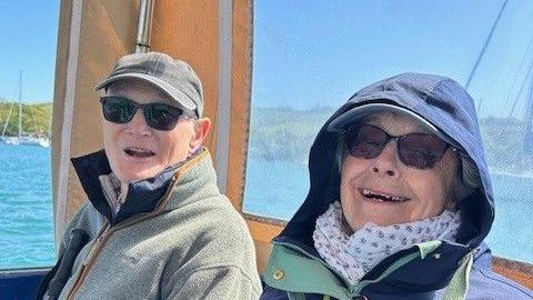 Ann Mallett and her husband sat on a boat. Both are wearing coats and sunglasses.