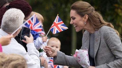 The Princess of Wales, wearing a grey suit and holding a small bunch of flowers, smiles as she meets a croud of people who are waving flags and taking photographs.