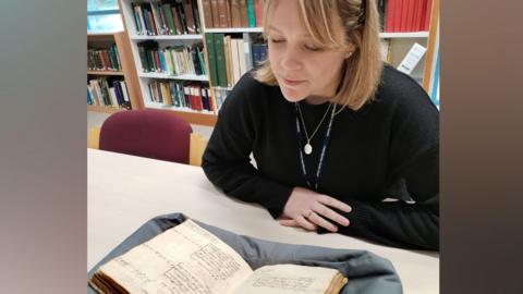Esther Bellamy looking at the terrier, which has been bound into a book. It resting on padded cloth on a white table top and is open to show two pages with black writing on them. She has a fair bob, is wearing a black top and is looking down at the book. behind her are rows of books in bookshelves.