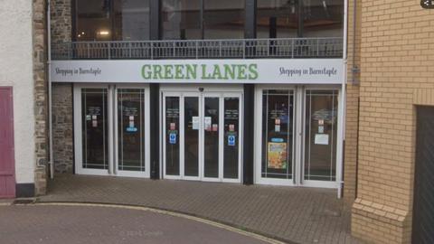 The outside of Green Lanes shopping centre in Barnstaple. It is a brick building with large glass windows and the words "Green Lanes" above the door. The entrance has large glass panels and, on the first floor, there are large glass windows.