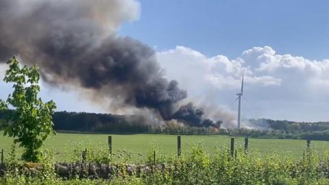 A fire next to a wind turbine in a green field with clouds of black smoke billowing into the sky 