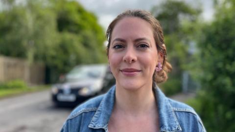 Ella Hodder wearing a blue denim jacket, she has brown hair and pink flowery earrings stood next to the country road with queuing traffic behind her