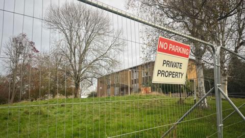 A metal barrier in front of the old police station building. A no parking private property is displayed on the fence. A grass bank leads up to the property.