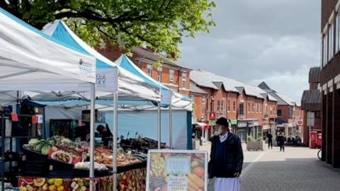 A man walking past a fruit and veg stall in a market on a high street