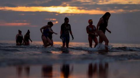 Seven women in swimming costumes wading pout of the sea at sunrise