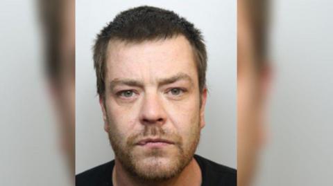 A mug shot of Stuart Hand who is a convicted paedophile. He is a white man with short brown hair and green eyes. He is wearing a black t-shirt. 