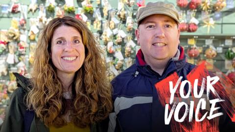 Wesley Thorne and his wife Toni are smiling and standing in front of a wall covered in Christmas decorations.