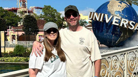 A woman with a white t-shirt, black shorts, white trainers, a grey baseball cap and sunglasses stood next to a man wearing a cream t-shirt, black shorts, white trainers, a black baseball cap and sunglasses against white railings, with the Universal Orlando resort in Florida pictured the background.