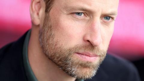 Close up shot of Prince William's face with stubble, he looks pensive and looks to the right of camera against a pink background