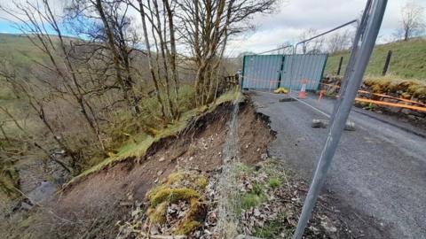 A single-track rural road is damaged on one side. A previous landslide has exposed mud on the hillside leading down to a body of water at the bottom of a valley. Metal fences and bright orange tape has been placed around the affected area.