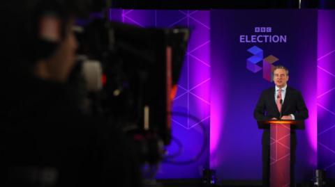 Nick Servini stood behind a podium for the BBC Ask the Leader debate, which is a purple background. There is a blurred cameraman in the foreground