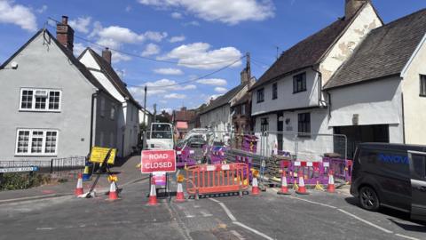 A High Street on a sunny day that has been closed off due to water works taking place. The road is lined by lots of houses and buildings.