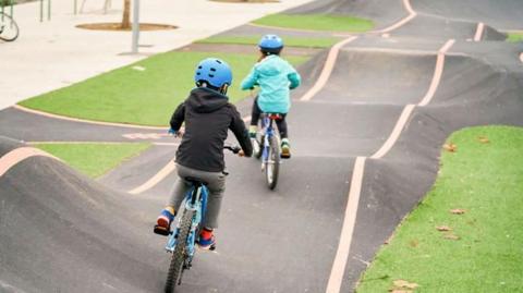 Two children ride bikes on a pump track in a park. The track has lines painted on either side and has bumps and dips. There are grass areas around the track.
