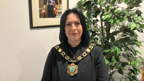 A woman with long dark hair and a dark top with a gold chain of office around her neck and a green plant behind her