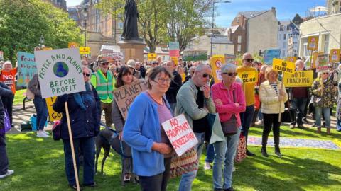 A crowd gathered in a park space holding signs opposing fracking, including messages about protecting the planet and prioritising people over profit.