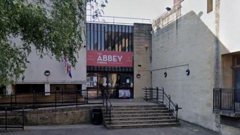 The front facade of abbey hall, including wide steps up to a large glass set of doors, with a red sign saying Abbey Cinema, in white writing, above it.