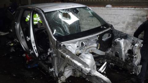 A stripped vehicle in a garage. The windscreen has been smashed and there is no engine or bonnet. 