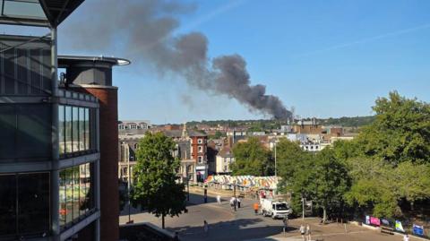 A view across a city centre. A plume of black smoke billows on the horizon against a blue sky. In the foreground are buildings, trees and the colourful canopies of Norwich market.