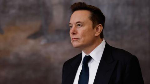 Elon Musk wearing a black suit, white shirt, and black tie looking into the distance