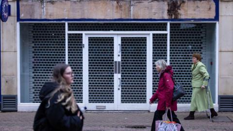 A group of women walking in different directions past an empty shop unit on a British High Street in winter