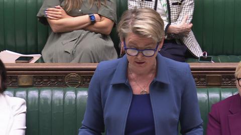 Yvette Cooper wearing a blue jacket and darker blue top and glasses, standing in the House of Commons