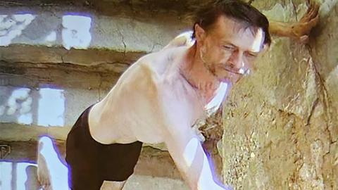 Pale-skinned man on light stone steps with beard, unkempt hair, black shorts, presses his hands against nearby stone wall