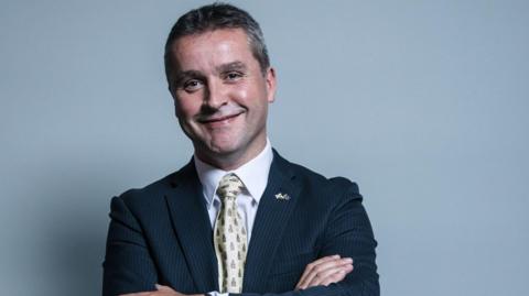 Angus MacNeil standing, arms folded, in front of a grey backdrop. He is smiling and wearing a blue suit with white shirt and yellow patterned tie.