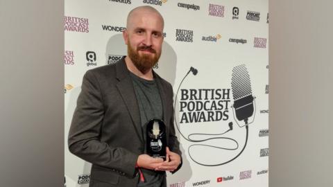 Matthew Butler, a bald man with a ginger beard in a dark grey blazer and a dark grey t-shirt holding a black and silver award. He's stood in front of an advertising wall. 