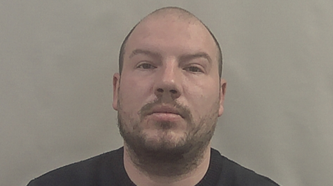 A police head shot of a man - Craig Hillman. He has a shaved head, short dark-coloured facial hair and is wearing a black t-shirt.