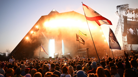 A stage at Glastonbury Festival 2022 is seen from the crowd. The sun is going down and the crowd is lit up in an orange light. There are a few flags held up by audience members.