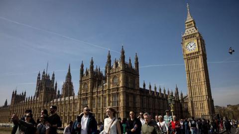 People walk across Westminster Bridge outside the Houses of Parliament in central London on a sunny day.