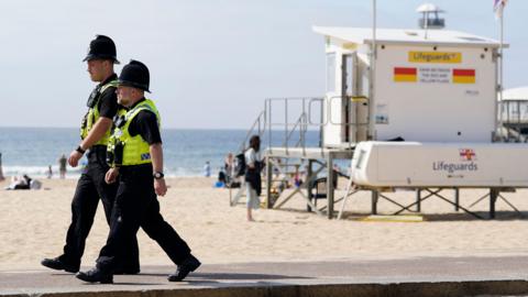Two police officers walking along the seafront in Bournemouth. There is a Lifeguards station behind them and several people on the beach in bright sunny weather