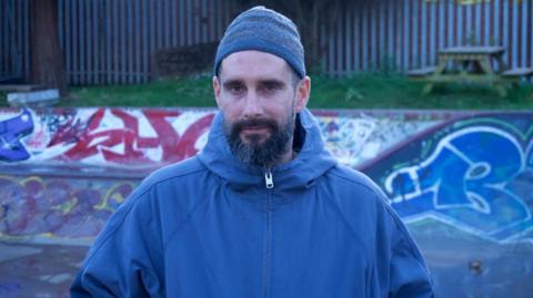 Rich Ward is wearing a grey beanie and a grey jacket. He is standing in front of skateboarding slopes filled with colourful graffiti 